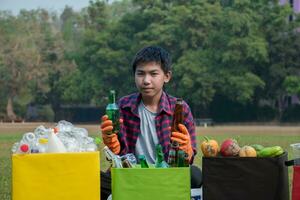 Aziatisch jongen in plaid overhemd zittend Aan speelplaats van school- met divers dozen van gescheiden kledingstukken of afval binnen, concept voor eco vrienden en globaal opwarming campagne, zacht en selectief focus. foto