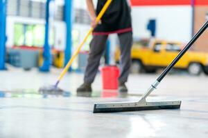 selecteer de focus dweil, onderhoud personeel Mens gebruik makend van een dweilen naar verwijderen water in de uniform schoonmaak de beschermend kleding van de nieuw epoxy verdieping in een leeg magazijn of auto onderhoud centrum. foto