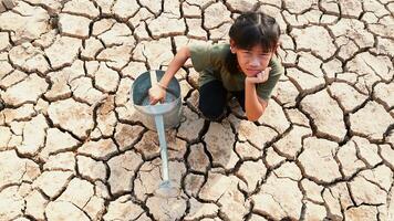 verdrietig weinig meisje zittend met een gieter kan Aan droog grond. water crisis, concept hoop en droogte foto