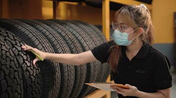 arbeider jong Azië vrouw vervelend gezicht medisch masker is controle kwaliteit van auto banden en controle de voorraad van auto banden in auto reparatie winkel op te slaan gedurende covid 19 epidemie. foto