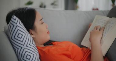 Aziatisch vrouw lezing een boek terwijl aan het liegen Aan de sofa foto