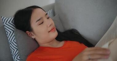 Aziatisch vrouw lezing een boek terwijl aan het liegen Aan de sofa foto