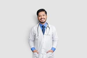 jonge mannelijke arts lachend met een stethoscoop en arm in zakken geïsoleerd op een witte achtergrond. zorgverzekeringsconcept. foto