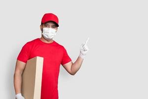 bezorger werknemer in rode dop leeg t-shirt uniform gezichtsmasker houd lege kartonnen doos geïsoleerd op witte achtergrond foto