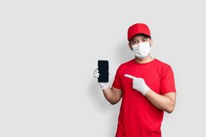 bezorger werknemer in rode dop leeg t-shirt uniform gezichtsmasker houd zwarte mobiele telefoon applicatie geïsoleerd op witte achtergrond foto