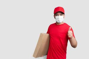 bezorger werknemer in rode dop leeg t-shirt uniform gezichtsmasker houd lege kartonnen doos met duimen omhoog geïsoleerd op witte achtergrond foto
