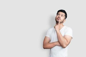Aziatische knappe man op wit t-shirt met twijfels terwijl hij opkijkt over geïsoleerde witte achtergrond foto
