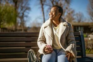 buitenshuis portret van gelukkig Afro-Amerikaans vrouw Aan zonnig dag. ze is zittend Aan bank in de straat en gebruik makend van smartphone. foto