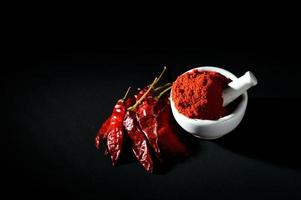 rode chili peper poeder in stamper met vijzel en rode chili pepers op zwarte achtergrond foto
