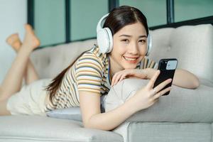 mooie aziatische vrouw liggend op de bank en luisterend naar muziek tijdens het gebruik van de telefoon foto