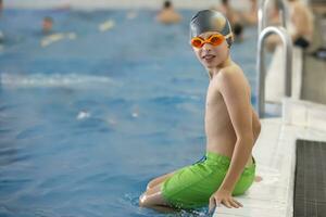 een jongen in een zwemmen pet en stofbril zit Aan de kant van de sport- zwembad. foto