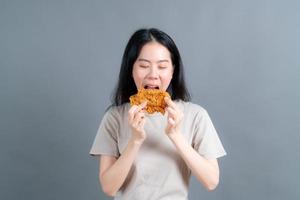 jonge aziatische vrouw die een trui draagt met een blij gezicht en geniet van het eten van gebakken kip op een grijze achtergrond