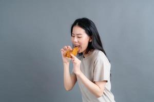 jonge aziatische vrouw die een trui draagt met een blij gezicht en geniet van het eten van gebakken kip op een grijze achtergrond foto