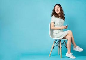 jong Aziatisch meisje zittend in een stoel met behulp van telefoon met verbaasde uitdrukking op de achtergrond foto