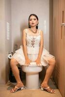 een Aziatisch vrouw in een wit jurk is zittend schrijlings een wit kast in een badkamer foto