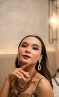 een Aziatisch vrouw met een goud ketting en oorbellen is poseren prachtig in een hotel kamer voordat avondeten foto