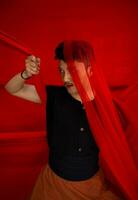een Aziatisch Mens in een zwart overhemd aan het bedekken zijn gezicht met een rood kleding met een beangstigend gelaats uitdrukking in voorkant foto