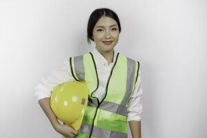 zelfverzekerd Aziatisch vrouw arbeid arbeider is glimlachen naar de camera, brengen haar veiligheid helm, vervelend geel veiligheid helm, groen hesje en uniform, geïsoleerd wit achtergrond. foto