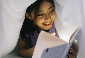 jong Aziatisch meisje ligt in een deken en gebruikt het licht van haar smartphone om een boek te lezen foto