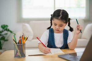 Aziatisch baby meisje glimlachen gebruik laptop of schrijven tekening kleur Aan Notitie boek studie online Aan hout tafel bureau in leven kamer Bij huis. onderwijs aan het leren online van huis concept. foto