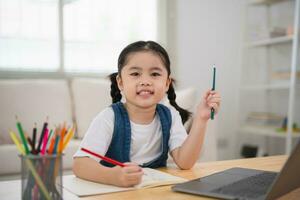 Aziatisch baby meisje glimlachen gebruik laptop of schrijven tekening kleur Aan Notitie boek studie online Aan hout tafel bureau in leven kamer Bij huis. onderwijs aan het leren online van huis concept. foto