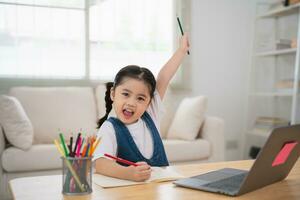 Aziatisch baby meisje glimlachen vrolijk en handen omhoog gebruik makend van laptop of schrijven tekening kleur Aan Notitie boek studie online Aan hout tafel bureau in leven kamer Bij huis. onderwijs aan het leren online van huis concept. foto