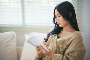 blij gelukkig Aziatisch vrouw glimlachen en lezing boek terwijl zittend Aan bankstel sofa in leven kamer Bij huis. Aziatisch Dames kom tot rust lezing boek glimlach Bij sofa in de huis. werkzaamheid hobby Bij huis concept. foto