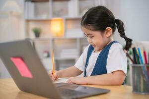 Aziatisch baby meisje glimlachen gebruik laptop of schrijven tekening kleur Aan Notitie boek studie online Aan hout tafel bureau in leven kamer Bij huis. onderwijs aan het leren online van huis concept. foto