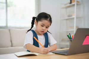 Aziatisch baby meisje glimlachen gebruik laptop of schrijven tekening kleur Aan Notitie boek studie online Aan hout tafel bureau in leven kamer Bij huis. onderwijs aan het leren online van huis concept. foto