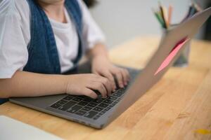 Aziatisch baby meisje glimlachen gebruik laptop of schrijven tekening kleur Aan Notitie boek studie online Aan hout tafel bureau in leven kamer Bij huis. onderwijs aan het leren online van huis concept. foto