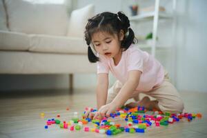 blij Aziatisch meisje gelukkig en glimlachen spelen kleurrijk Lego speelgoed, zittend Aan de leven kamer vloer, creatief spelen met Lego, gebouw kleurrijk structuren creativiteit stel je voor. aan het leren onderwijs. foto