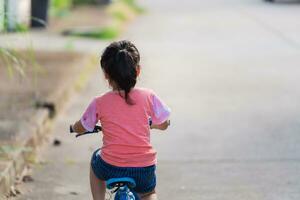 achterkant van Aziatisch kleuter meisje kind aan het leren naar rijden fiets in zonnig zomer dag, kind wielersport Bij park, baby sport concept foto