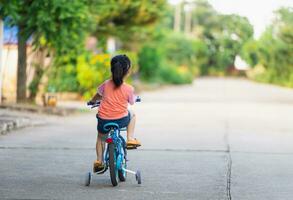 achterkant van Aziatisch kleuter meisje kind aan het leren naar rijden fiets in zonnig zomer dag, kind wielersport Bij park, baby sport concept foto
