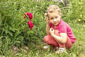 het kleine meisje is in gedachten verzonken, zittend bij het bloembed in de tuin van roze pioenrozen foto