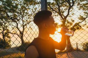 zwart Mens aan het doen sport- in ochtend, drinken water Aan basketbal rechtbank Aan zonsopkomst foto