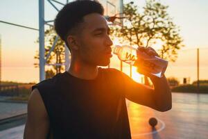 zwart Mens aan het doen sport- in ochtend, drinken water Aan basketbal rechtbank Aan zonsopkomst foto