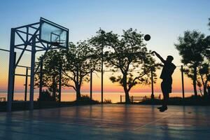 koel zwart Mens aan het doen sport, spelen basketbal Aan zonsopkomst, jumping silhouet foto