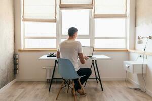 jong knap glimlachen Mens in gewoontjes kleding zittend Bij tafel werken Aan laptop foto