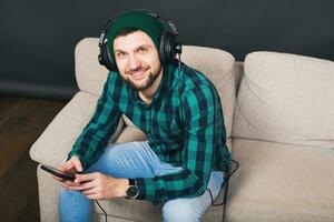 jong hipster knap gebaard Mens zittend Aan een bankstel Bij huis, luisteren naar muziek- foto