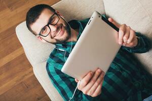 jong hipster knap Mens aan het liegen Aan bankstel Bij huis Holding tablet foto