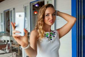 aantrekkelijk vrouw in wit jurk in zomer cafe in zonnebril gebruik makend van telefoon nemen foto