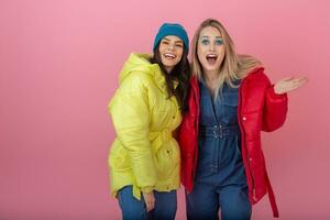 twee aantrekkelijk meisje vrienden actief Dames poseren Aan roze achtergrond in kleurrijk winter naar beneden jasje van helder rood en geel kleur hebben pret samen, warm jas mode tendens, vriendelijke groet lachend foto