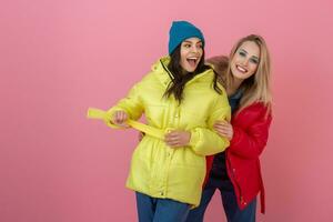 twee opgewonden aantrekkelijk meisje vrienden actief Dames poseren Aan roze achtergrond in kleurrijk winter naar beneden jasje van helder rood en geel kleur hebben pret samen, warm jas mode neiging foto