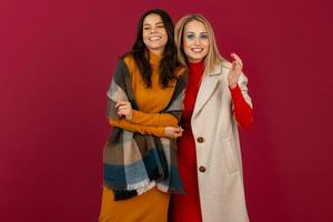 twee glimlachen aantrekkelijk elegant Dames in herfst winter mode jurk en jas poseren geïsoleerd Aan rood studio achtergrond foto