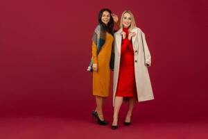 twee elegant Dames in herfst winter mode jurk en jas poseren geïsoleerd vol hoogte Aan rood studio achtergrond foto