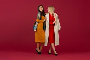 twee elegant Dames in herfst winter mode jurk en jas poseren geïsoleerd vol hoogte Aan rood studio achtergrond foto