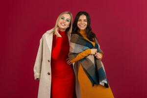 twee glimlachen aantrekkelijk elegant Dames in herfst winter mode jurk en jas poseren geïsoleerd Aan rood studio achtergrond foto