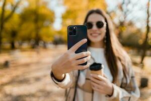 aantrekkelijk jong vrouw wandelen in herfst vervelend jasje gebruik makend van telefoon foto