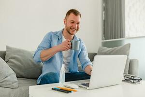 knap glimlachen Mens in overhemd zittend ontspannen Aan sofa Bij huis Bij tafel werken online Aan laptop foto