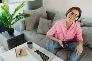 vrouw in roze overhemd zittend ontspannen Aan sofa Bij huis Bij tafel werken online Aan laptop foto